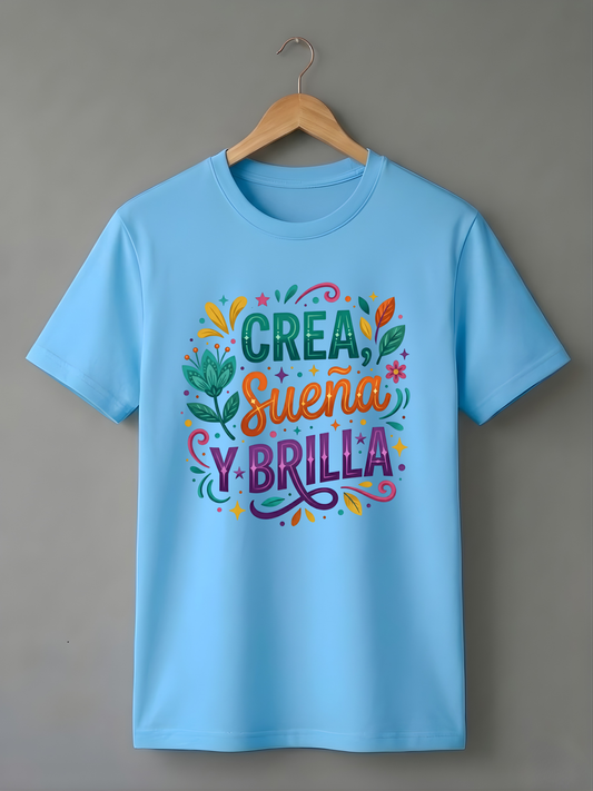 Vista frontal de camiseta azul cielo en percha. Presenta el diseño artístico "Crea, Sueña y Brilla" con detalles botánicos y colores vivos.